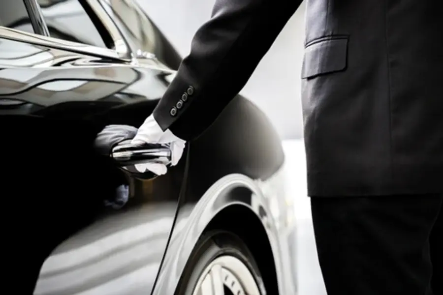 A driver in uniform holds the door of a black luxury sedan, preparing to open it, symbolizing luxury corporate transportation services.
